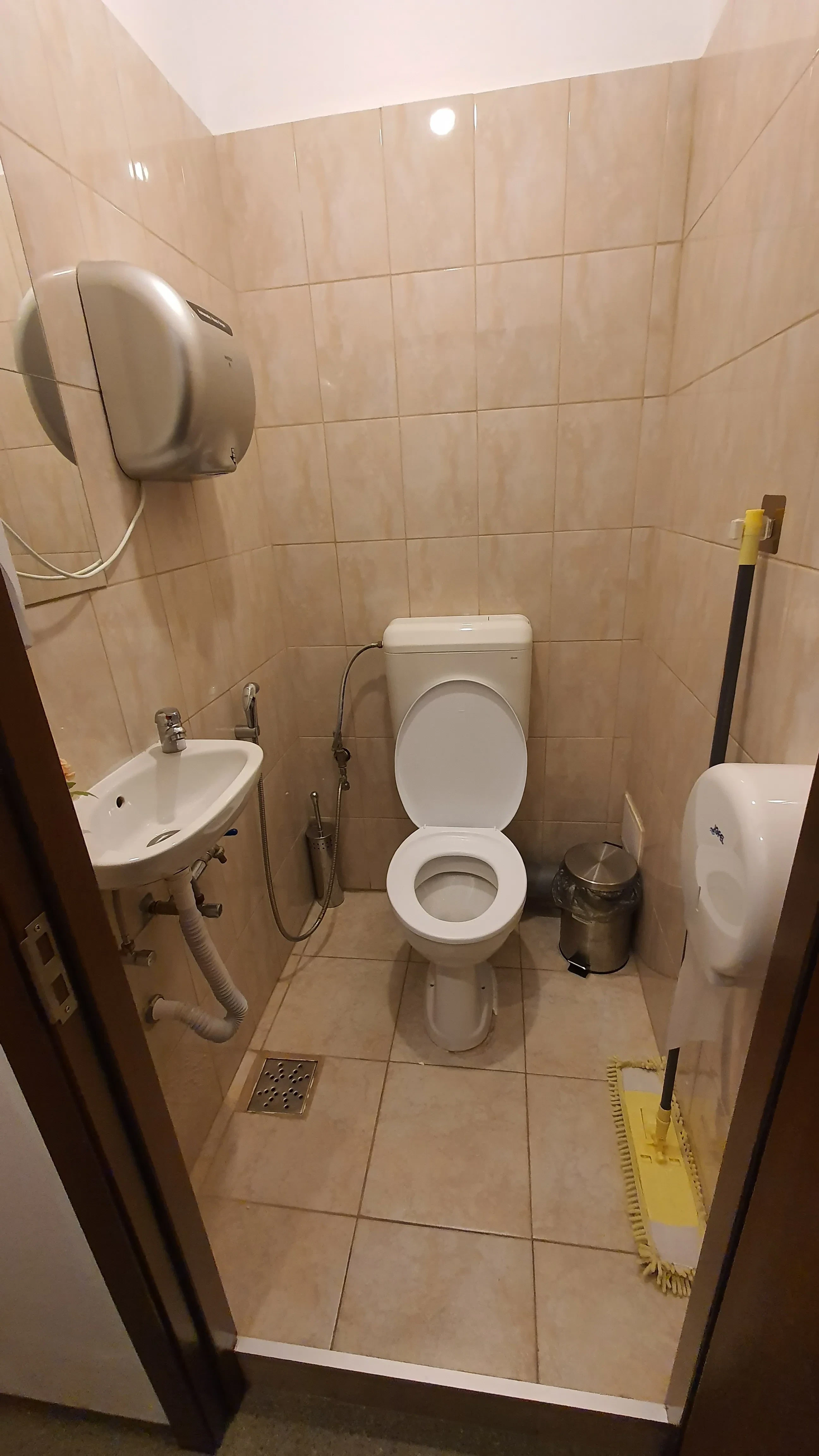 Salle de bain partagée au Popcorn Hostel : toilette, lavabo, distributeur de serviettes en papier et carrelage beige.
