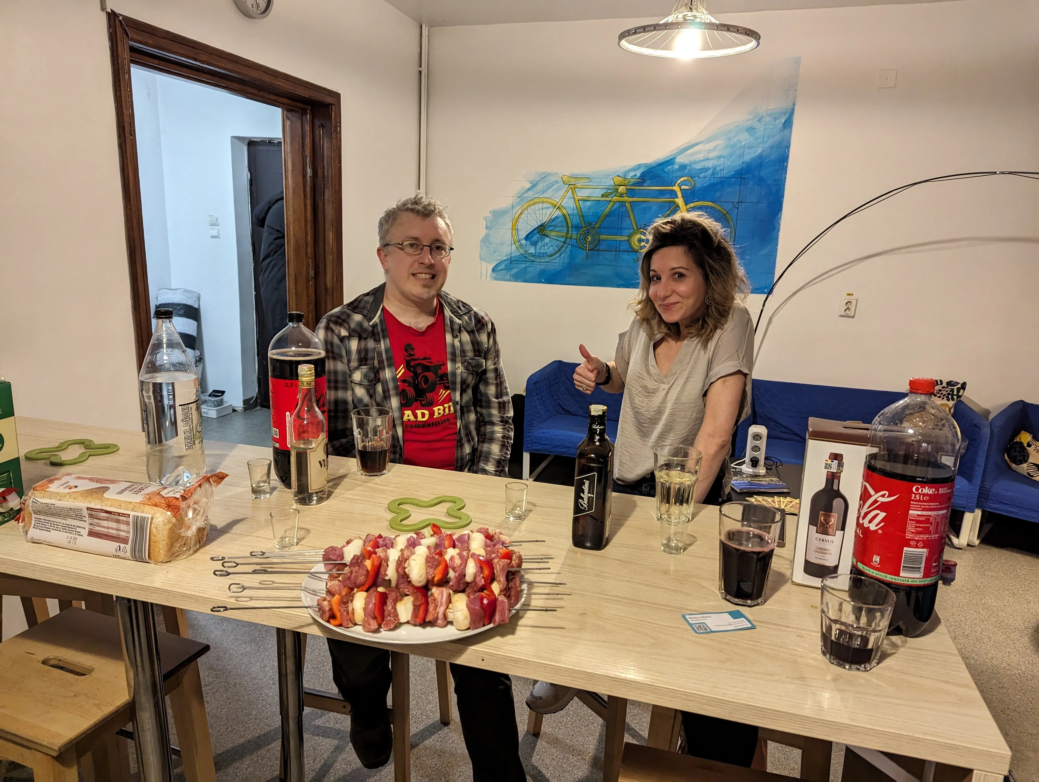 Deux voyageurs à la grande table en bois dans l'espace commun du Popcorn Hostel, avec brochettes et boissons sur la table.
