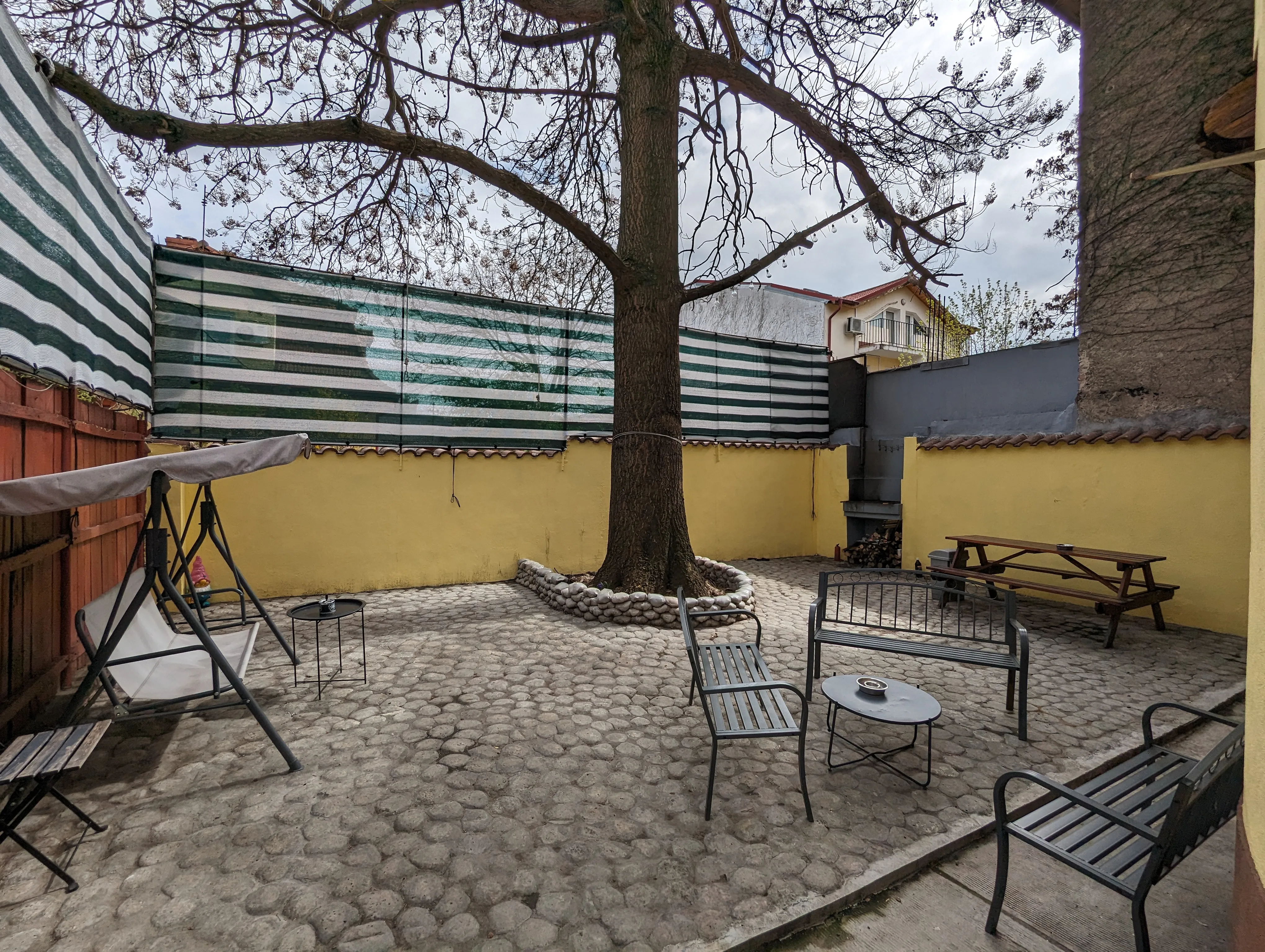 Cour privée du Popcorn Hostel : grand arbre, table de pique-nique, four maçonné et murs jaunes.