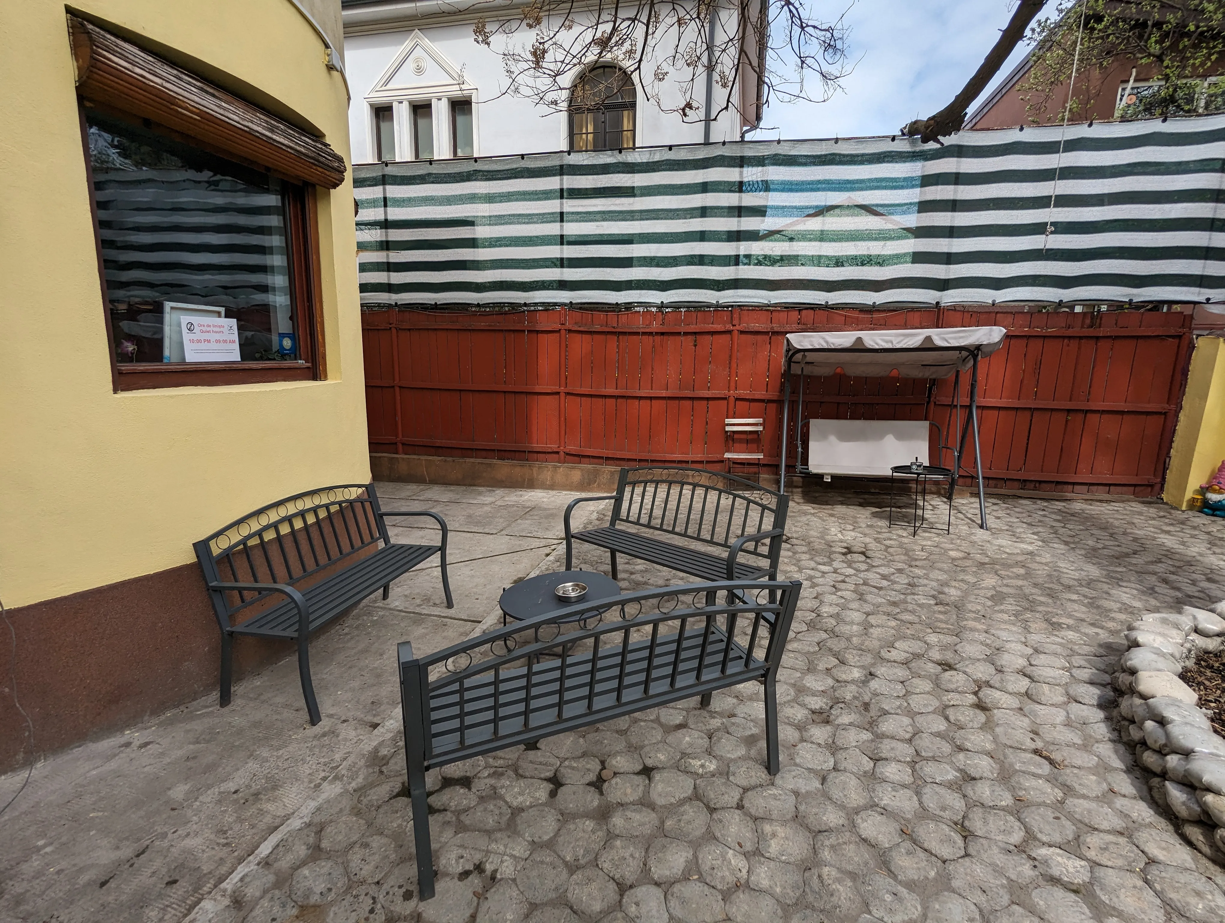Trois bancs en métal disposés en U près de l'entrée du Popcorn Hostel, sous une bâche à rayures vert-blanc.
