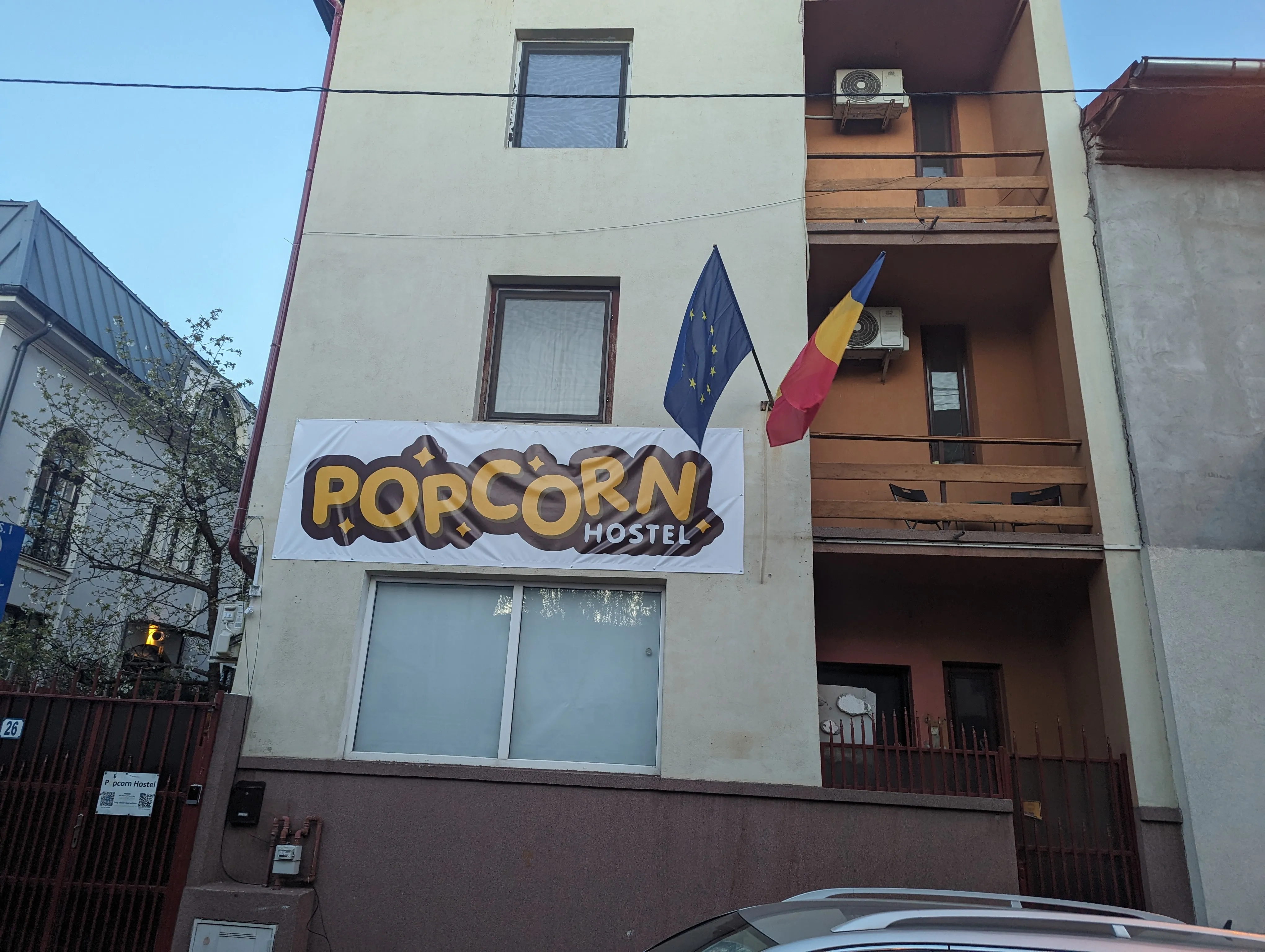 Façade du Popcorn Hostel au crépuscule, avec drapeau UE et drapeau roumain au balcon près de la banderole orange du logo.