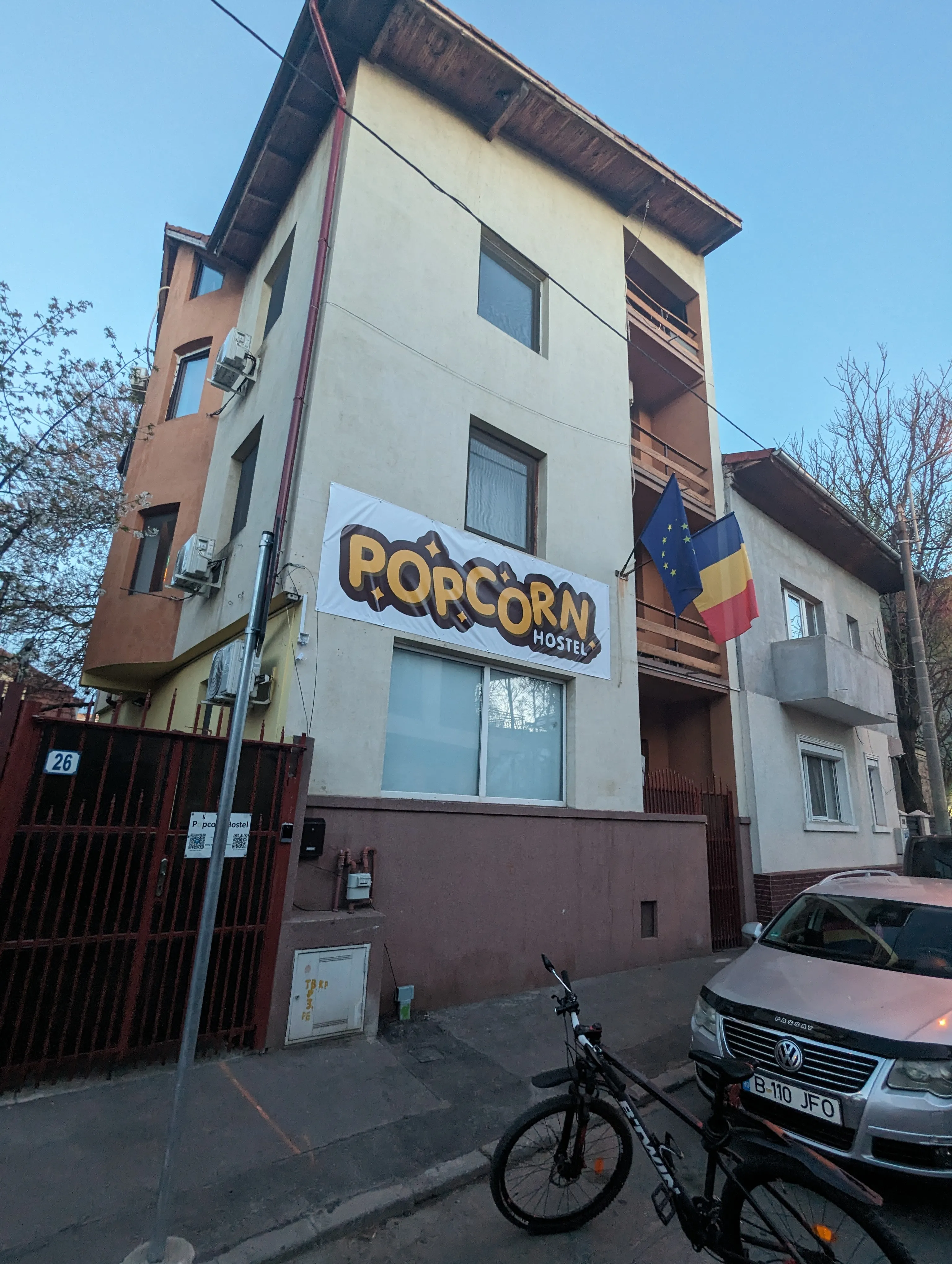Rue Ing. Pisoni 26 au crépuscule, avec le Popcorn Hostel à gauche et un vélo garé devant le portail rouge.