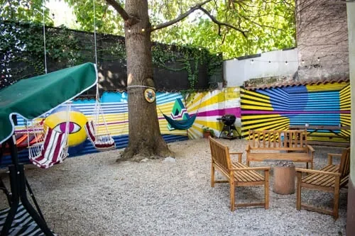 Cortile del Popcorn Hostel con albero centrale, altalene, amache, panche in legno e murales con occhi gialli sul recinto.