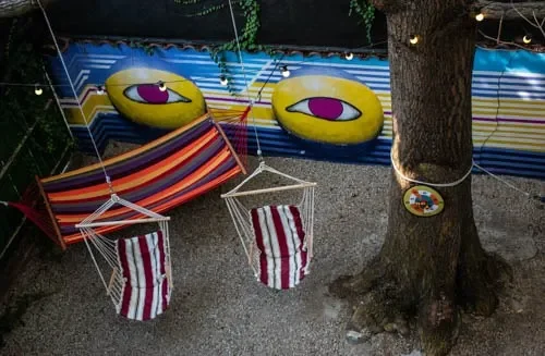 Amaca a strisce e due altalene sospese all'albero del cortile del Popcorn Hostel, con luci e murales urbano.