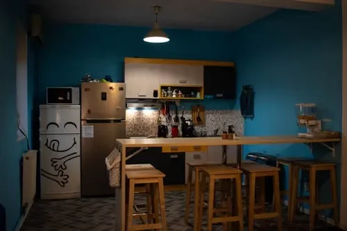 Cucina condivisa di sera al Popcorn Hostel: pareti blu, frigorifero con adesivo sorridente e lungo bancone colazione.