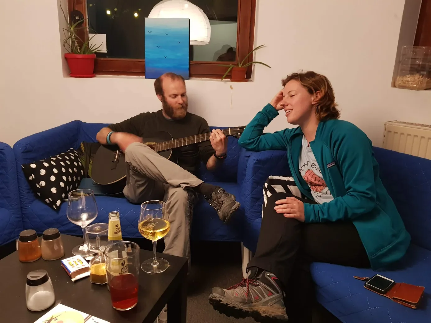 Due viaggiatori conversano con la chitarra sul divano blu del lounge al Popcorn Hostel, calici di vino sul tavolo.