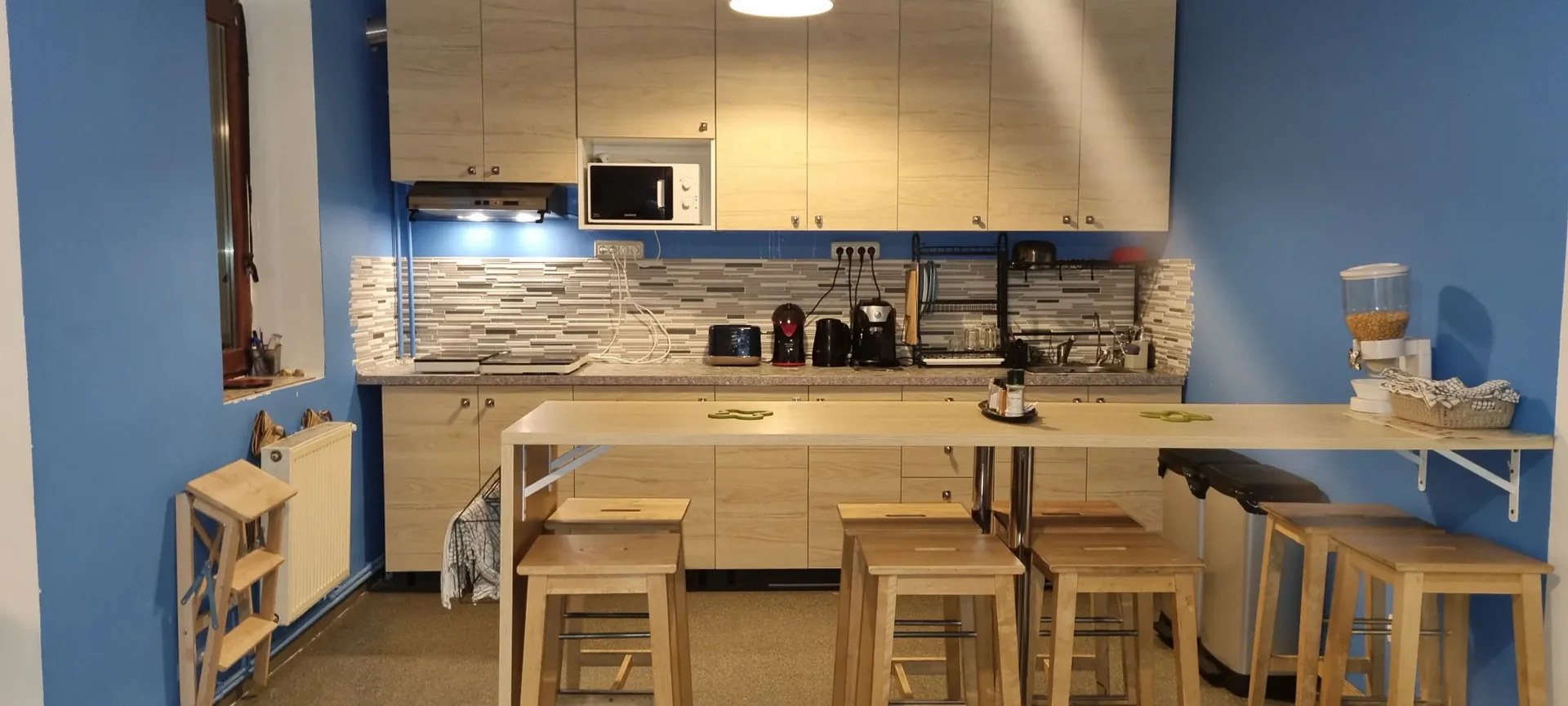Cucina condivisa al Popcorn Hostel con bancone colazione, alti sgabelli in legno e pareti blu.