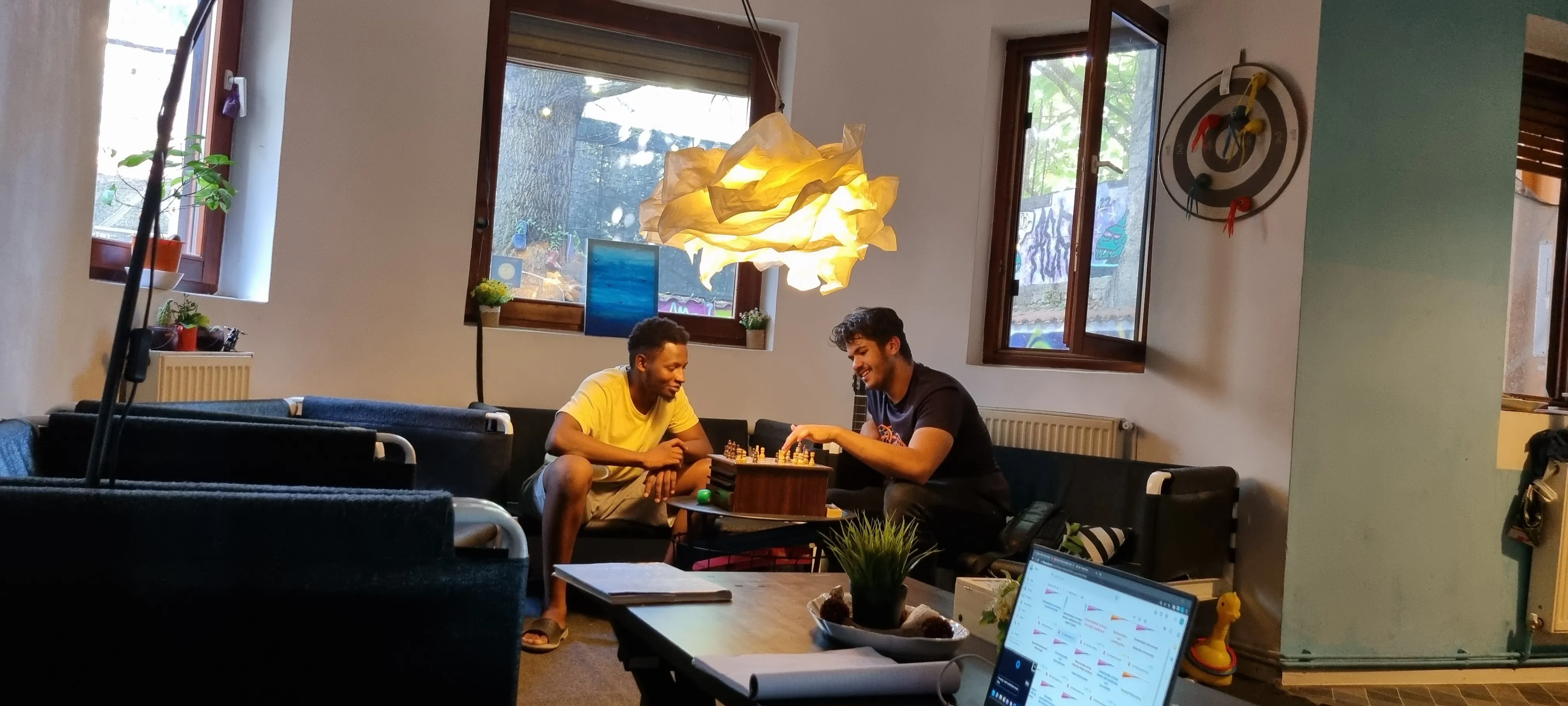 Deux voyageurs jouent aux échecs à la table basse du salon du Popcorn Hostel, lumière naturelle des fenêtres sur la cour.