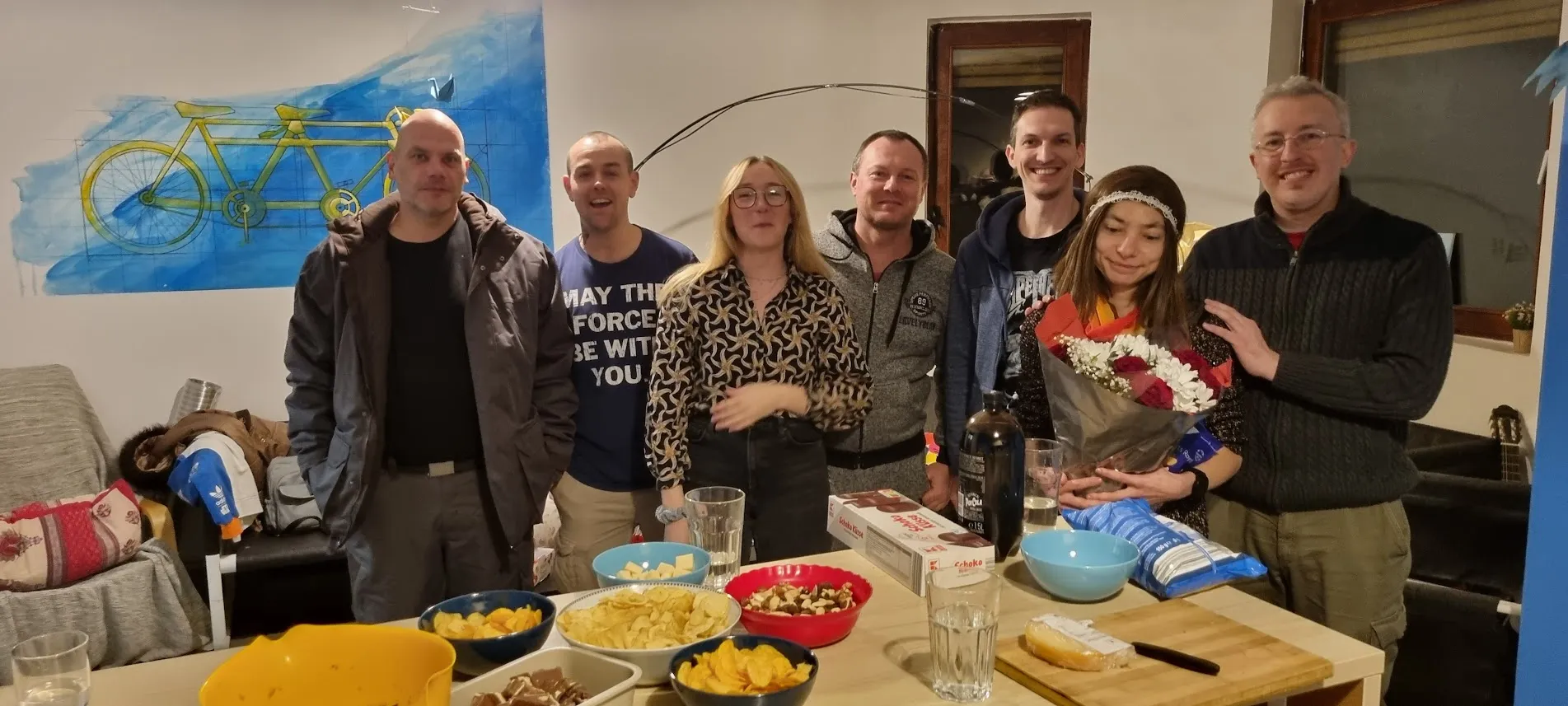 Groupe de six voyageurs souriants à la table d'apéritif du salon du Popcorn Hostel, sous la lampe aux pétales de papier.