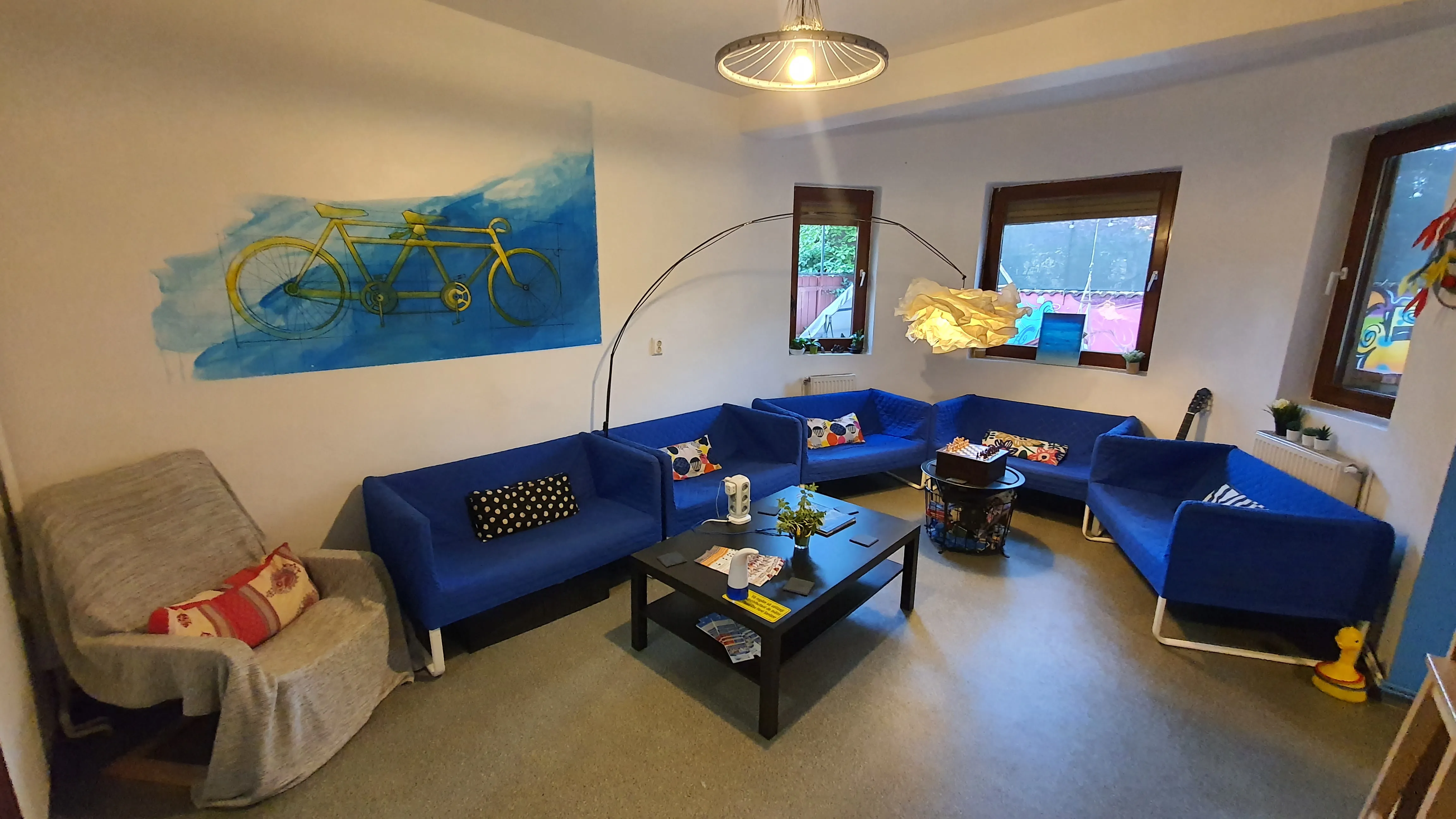 Salon commun du Popcorn Hostel : canapés bleus, lampe arc, toile au tandem jaune et fauteuil de lecture.