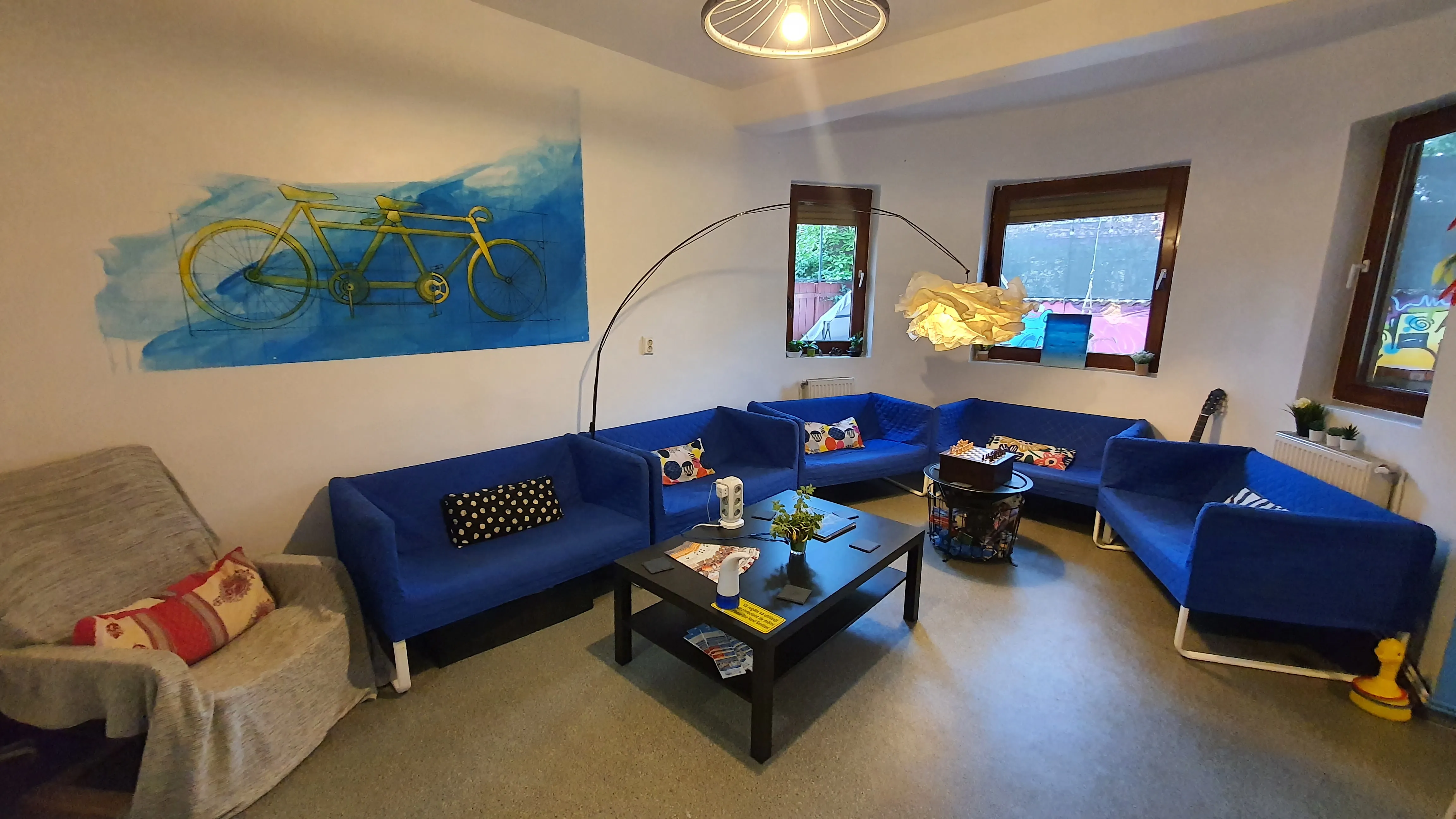 Salon du Popcorn Hostel vu du bout de la table commune, avec fauteuil et plaid gris dans le coin lecture.