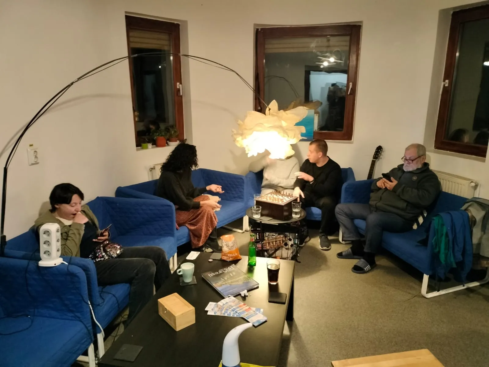 Cinq voyageurs détendus le soir sur les canapés bleus du salon du Popcorn Hostel, avec un jeu d'échecs sur la table.